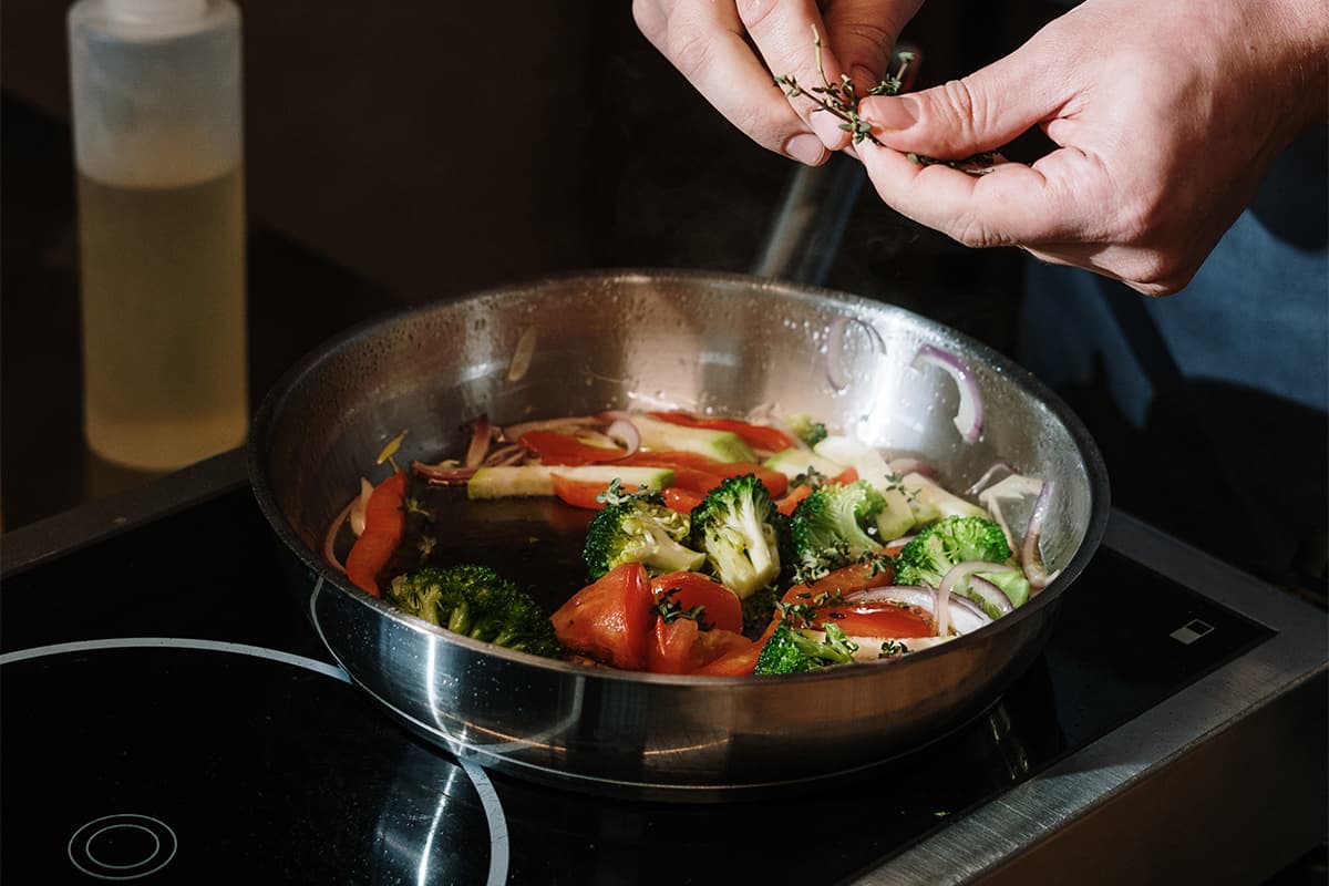 hands holding herbs above an induction cooktop filled with vegetables