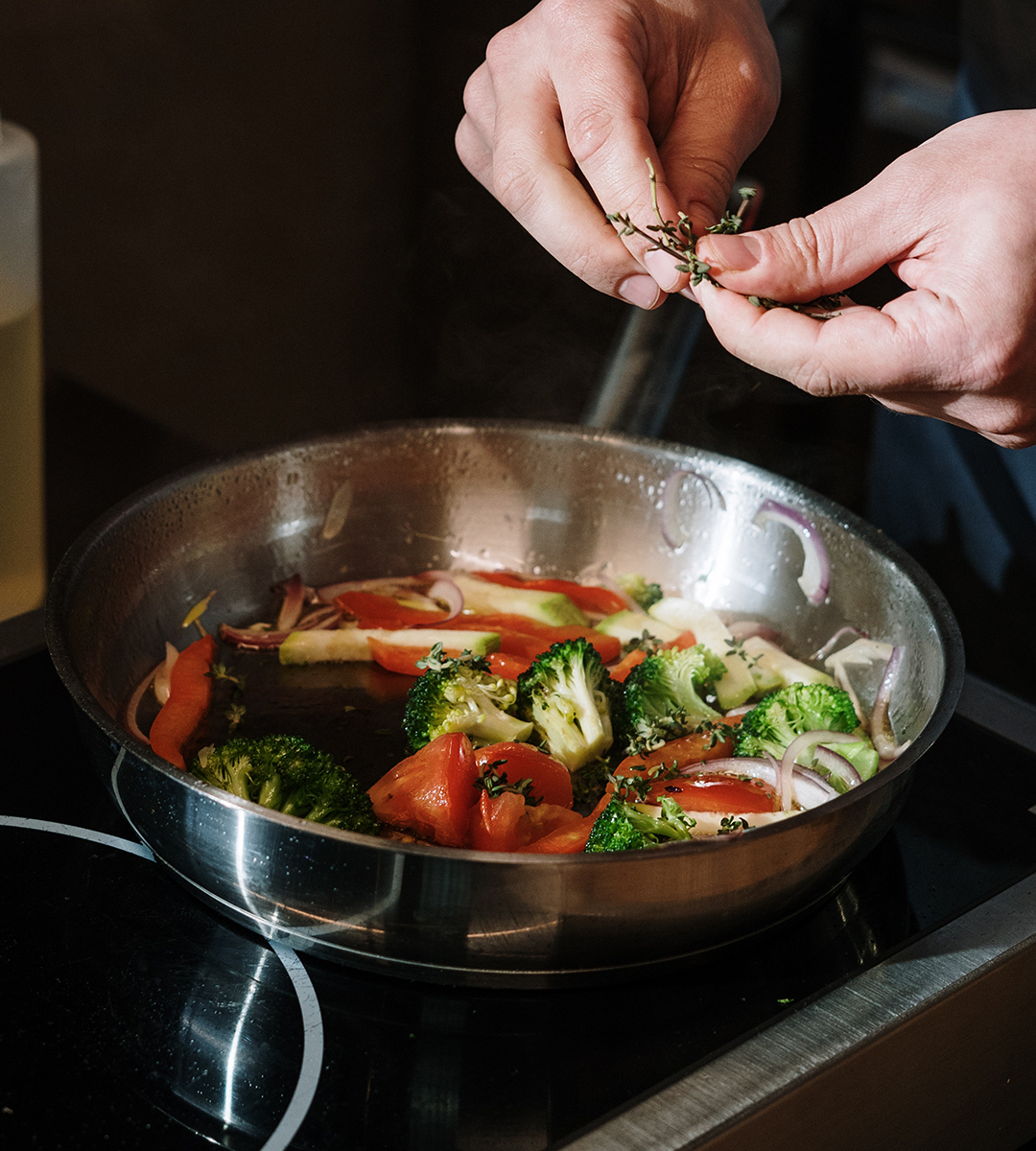 Hands holding herbs over an induction cooktop with vegetables