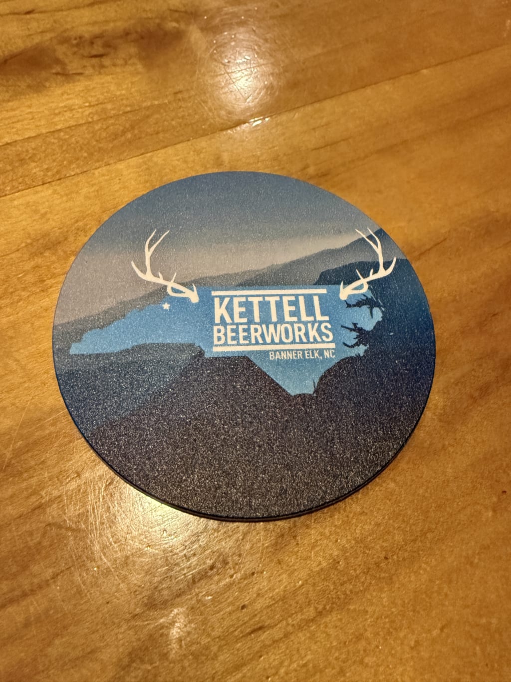 Round Kettell Beerworks coaster with North Carolina map and antler design on wooden table.