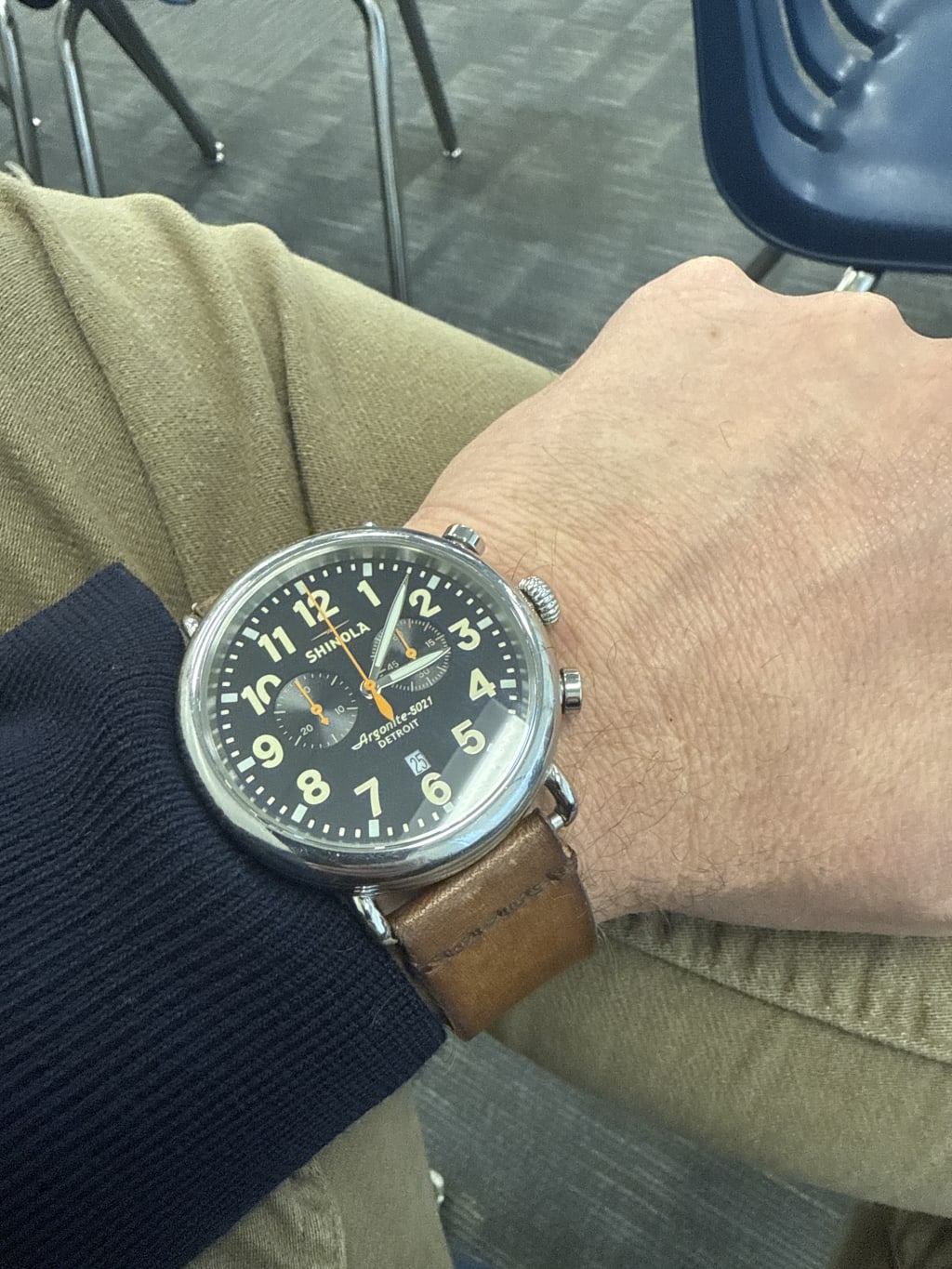 A person wearing a Shinola wristwatch with a black dial and brown leather strap.