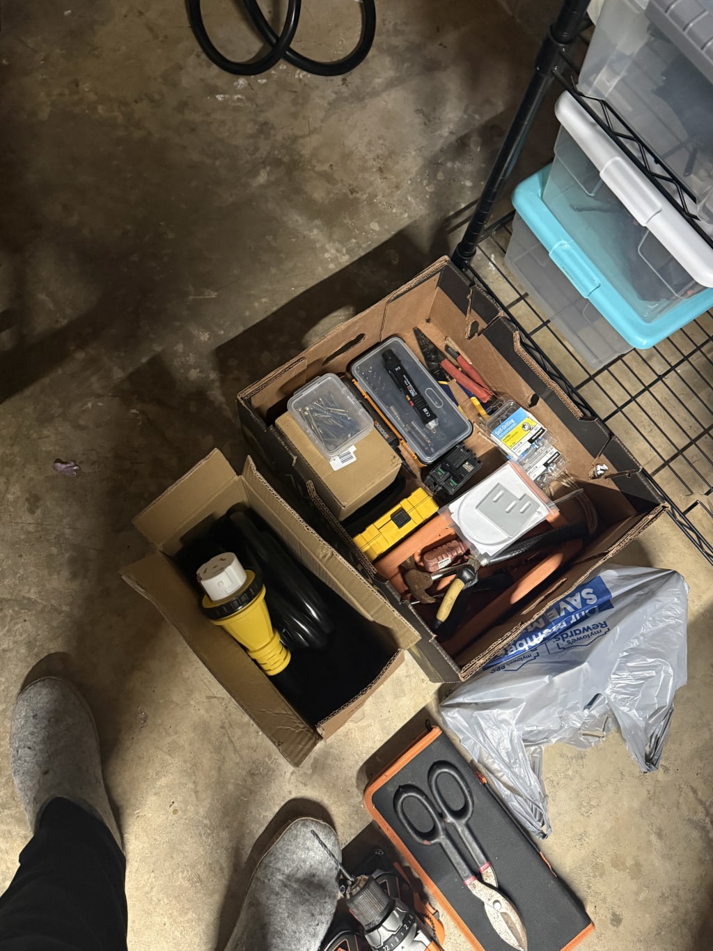 Two cardboard boxes with tools and an extension cord on a concrete floor near shelving.