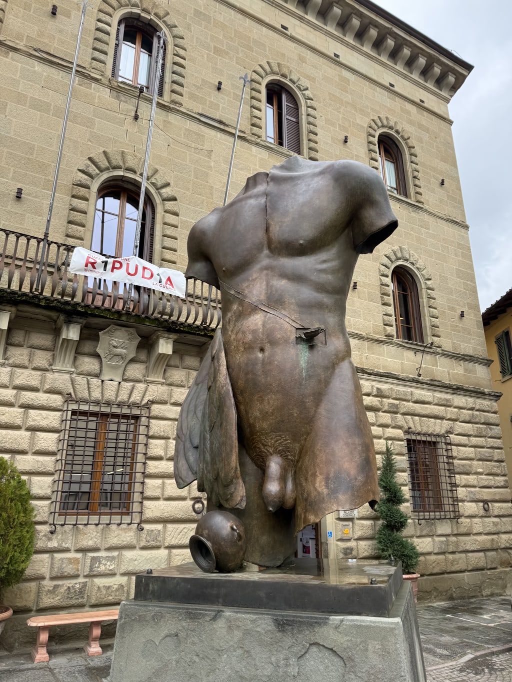Large bronze torso sculpture with missing head and arms in front of stone building.
