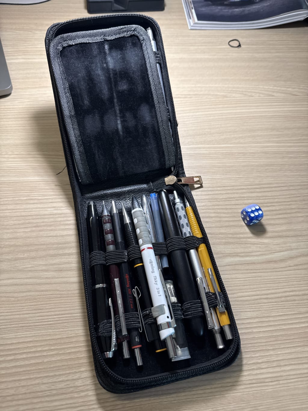 A black zippered pencil case containing eight pens and pencils next to a blue die.