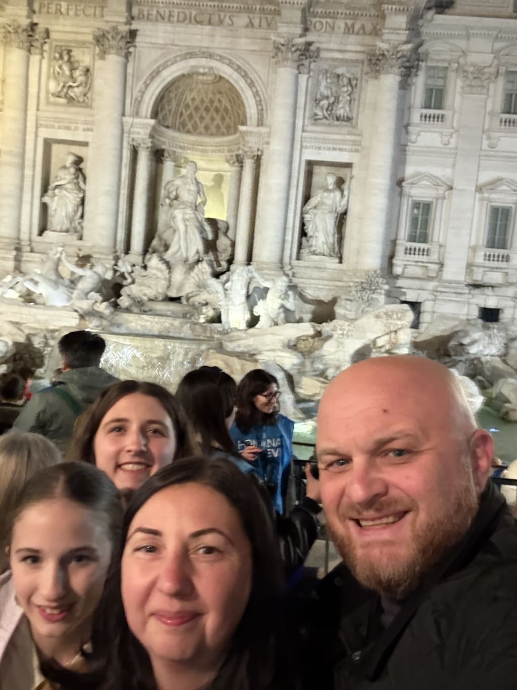 Busy crowds and us smiling and posing in front of the illuminated Trevi Fountain at night.