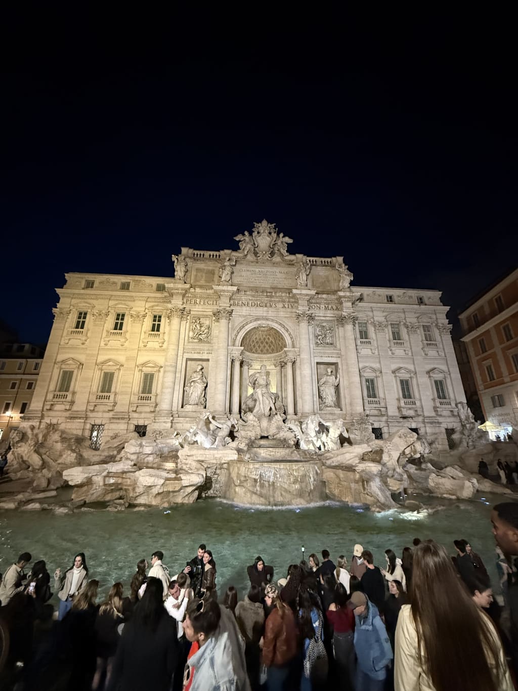 The illuminated Trevi Fountain in Rome at night with a crowd of people gathered.