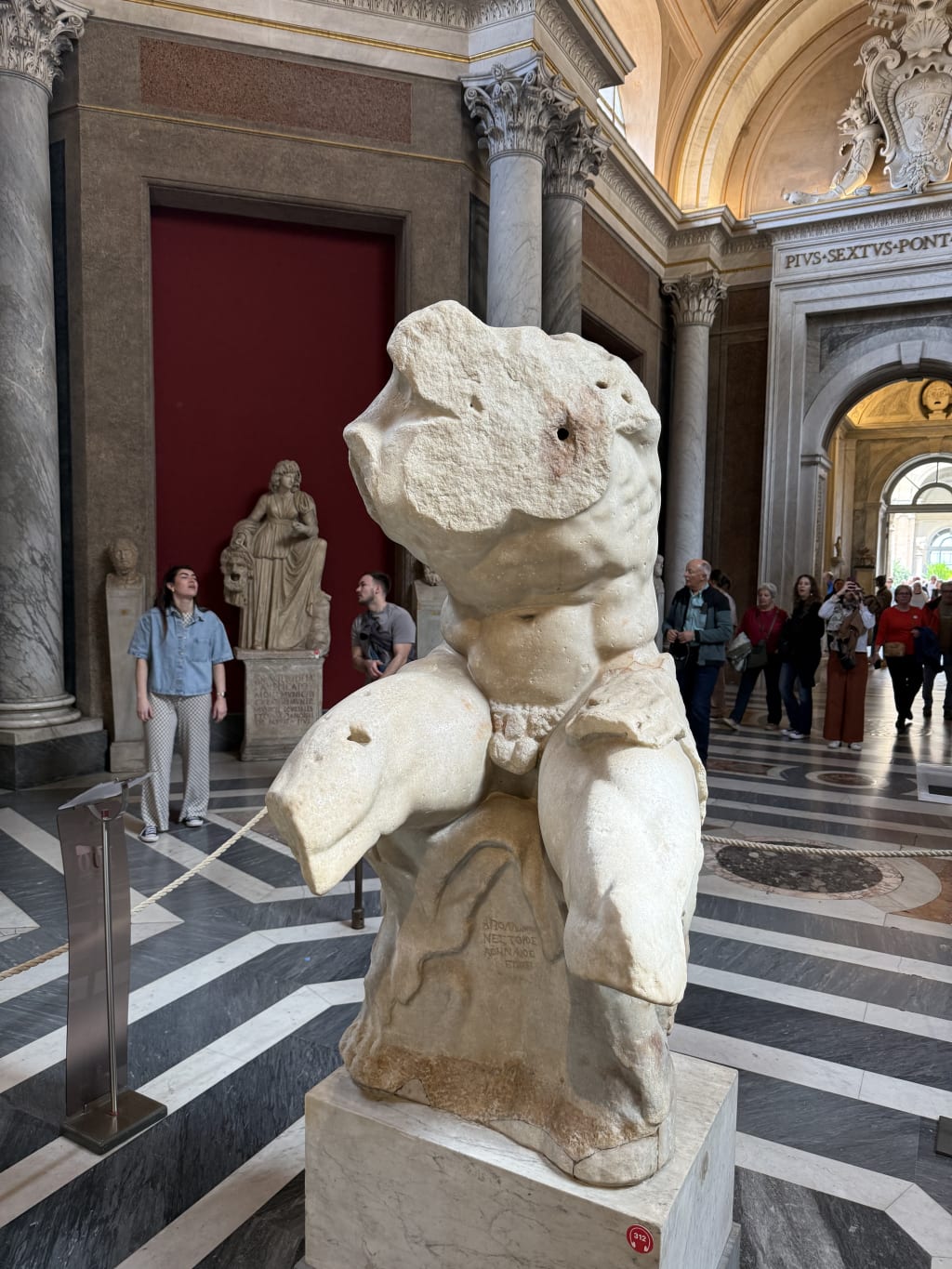 Ancient marble statue of a muscular male torso displayed in a museum with visitors.