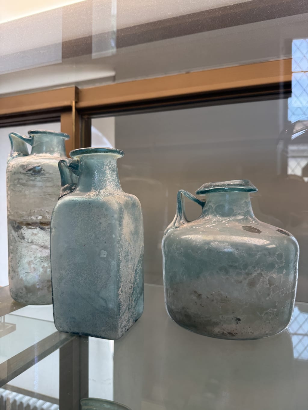 Three ancient, weathered blue-green glass bottles with handles displayed on glass shelf.