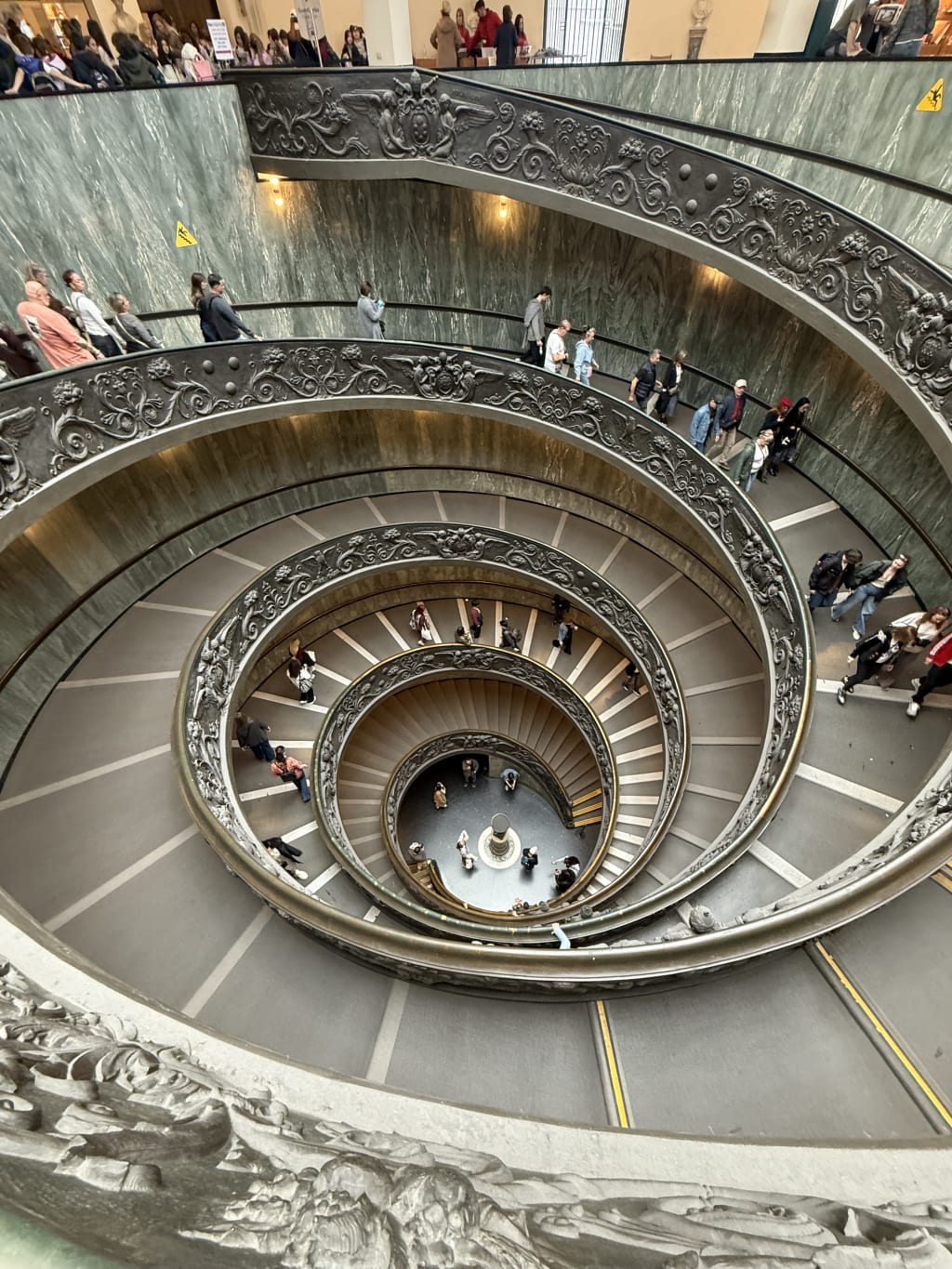 Looking down at a crowded, ornate spiral staircase with sculpted railings in the Vatican museum.