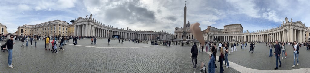 A panoramic view of St. Peter's Square filled with tourists on a partly cloudy day.
