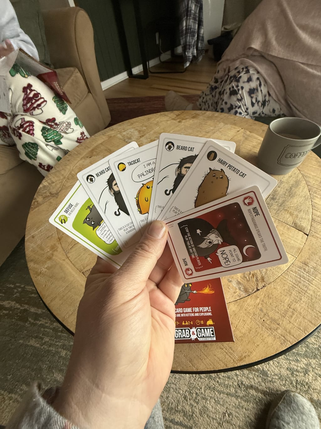 A person holding a hand of "Exploding Kittens" cards over a wooden table.