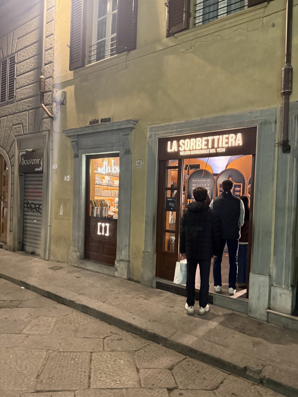 People are standing outside La Sorbettiera gelato shop at night on a stone-paved street.