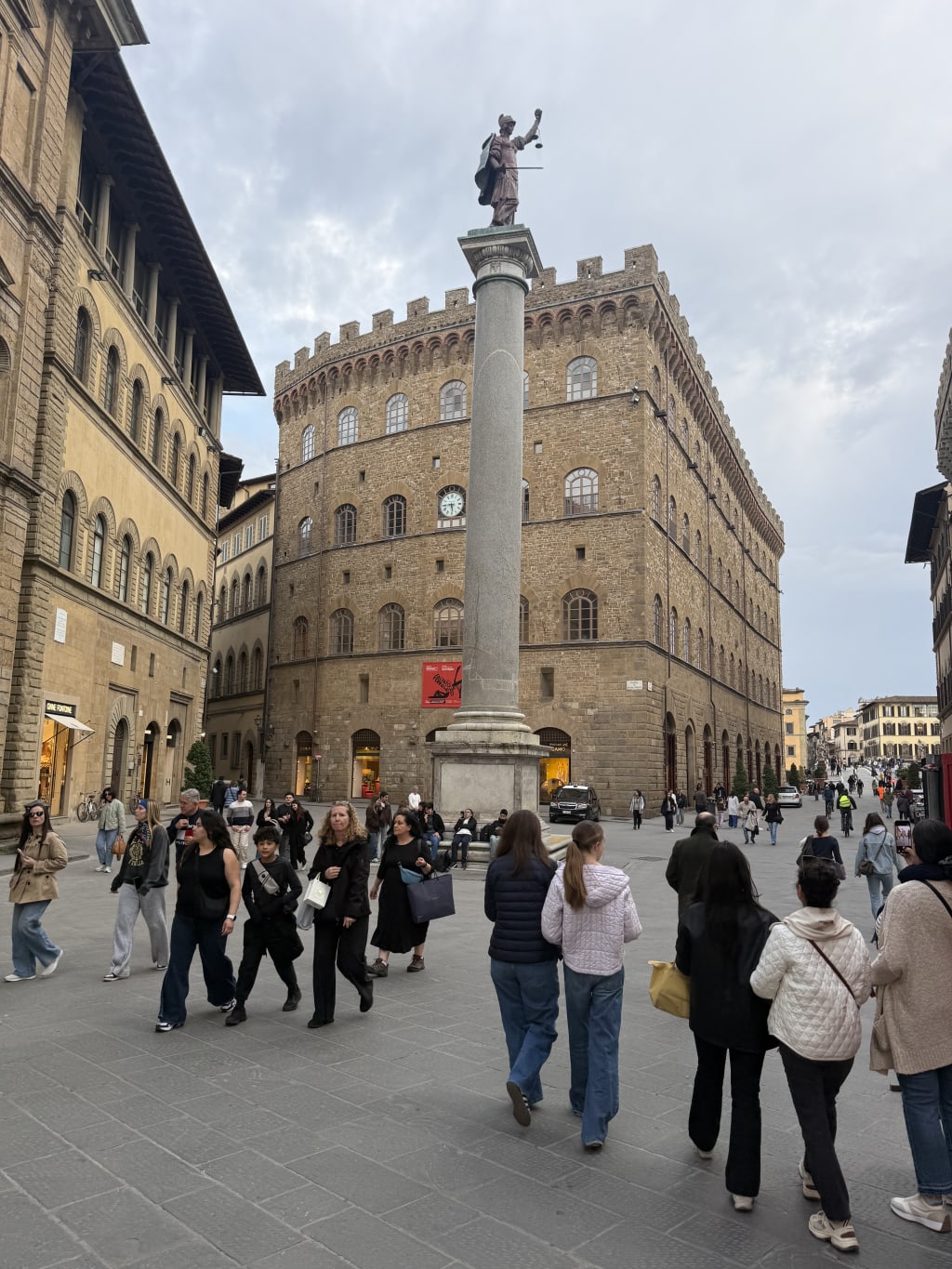 A busy square in Florence, Italy, featuring the Column of Justice and historic buildings.