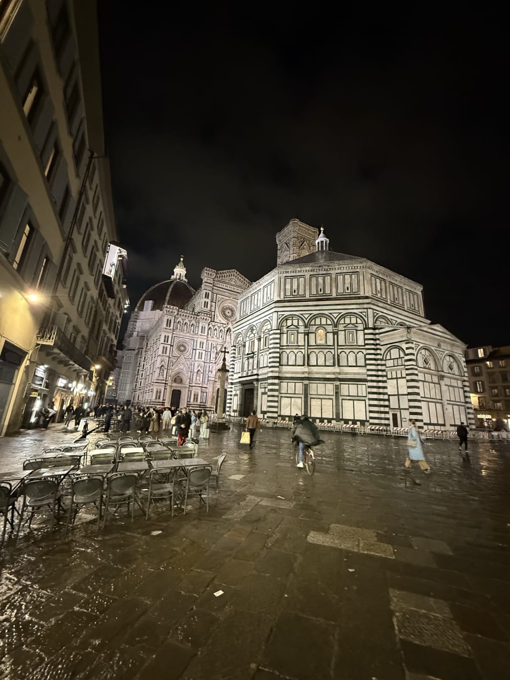 Nighttime view of Florence Cathedral and Baptistery with outdoor cafe tables and pedestrians.