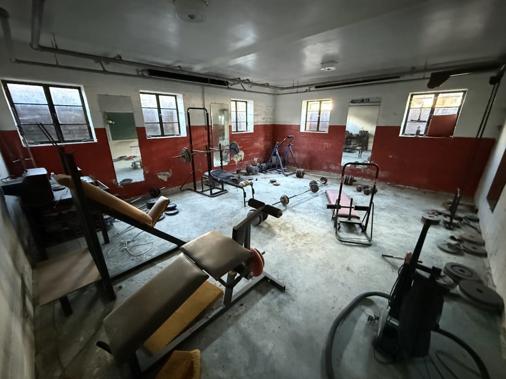A dimly lit, worn-out gym with weights, exercise equipment, and cracked red and white walls.
