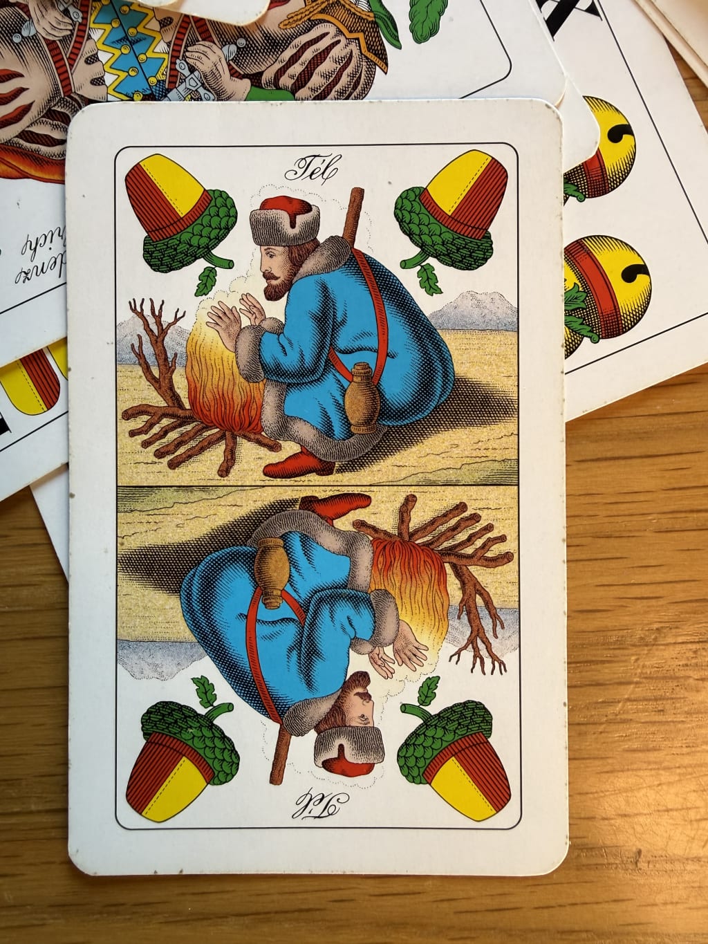 A playing card depicting a man warming hands by a fire, adorned with acorn symbols.  This is the ace of ‘acorns’.