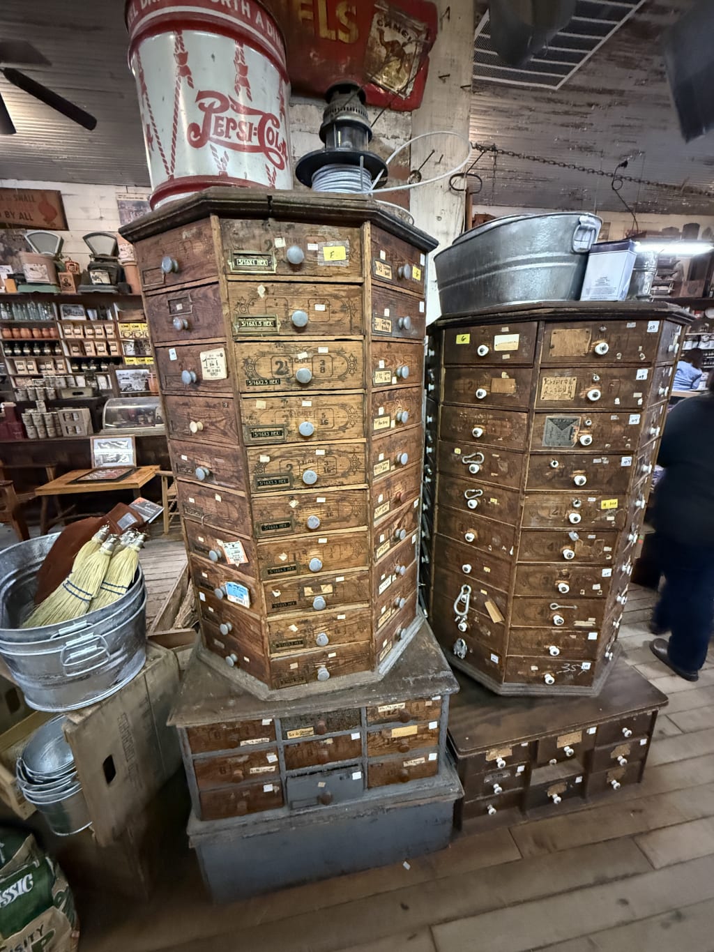 Two vintage multi-drawer wooden cabinets in a rustic store with metal buckets and brooms nearby.