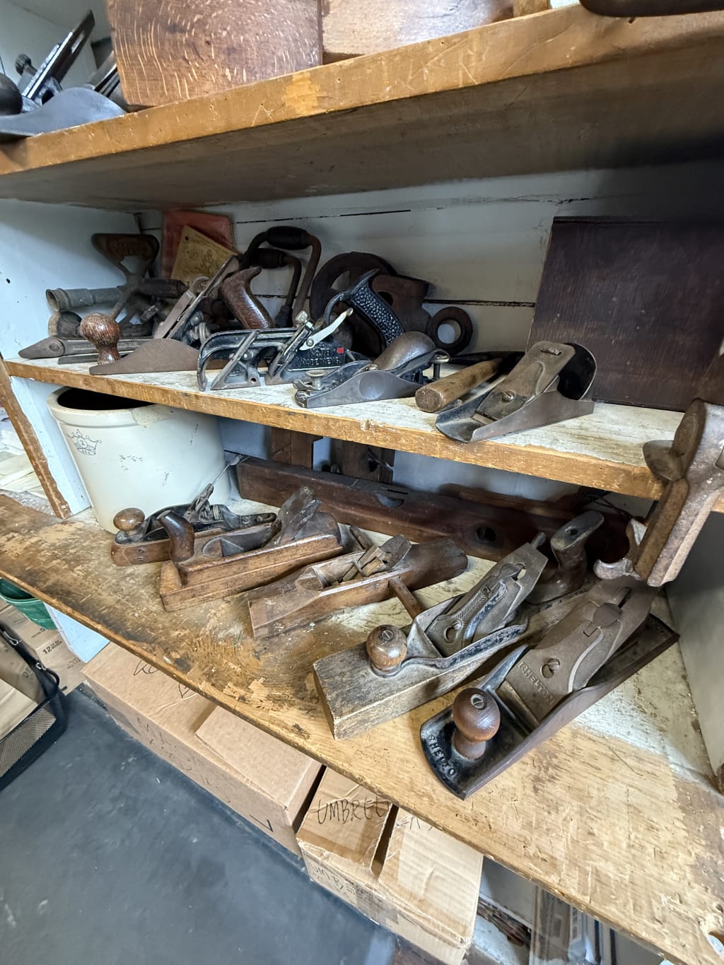 A collection of vintage hand planes and woodworking tools on wooden shelves in a workshop.