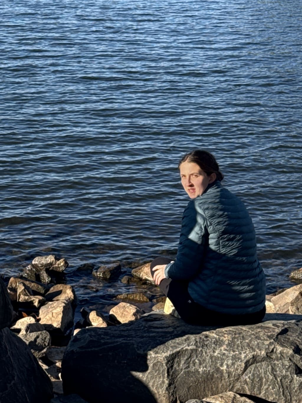 A person wearing a blue jacket sits on rocks by a body of water.