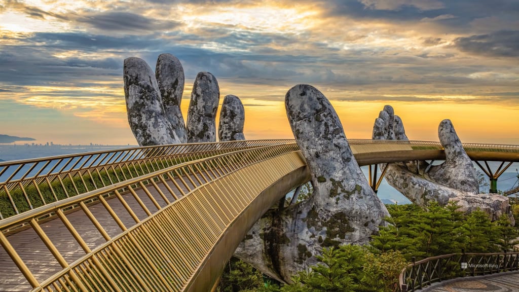 A golden pedestrian bridge held up by giant stone hands under a vibrant sunset sky.