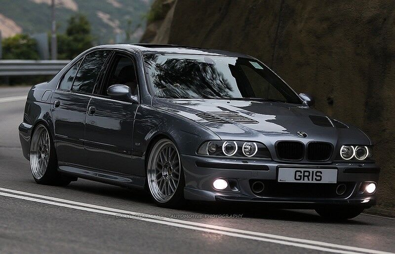 A dark gray BMW E39 sedan with custom wheels and illuminated halo headlights.