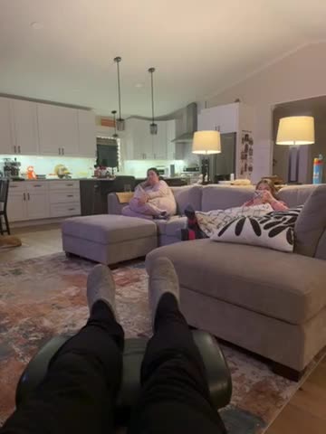 Two people are sitting on a couch in a well-lit, modern kitchen and living room.