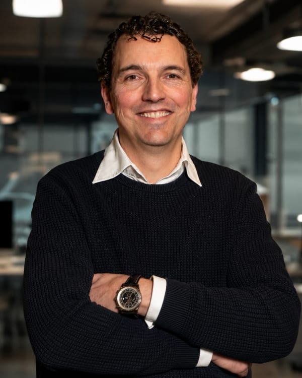 Smiling man with curly hair and a dark sweater, arms crossed in a modern office setting.