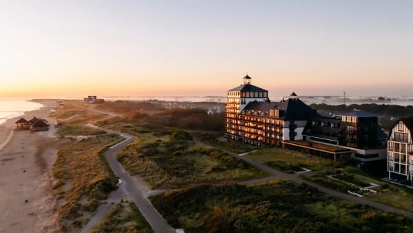 Sunset over a coastal resort with a large multi-story hotel by the dunes and a winding path along the beach.