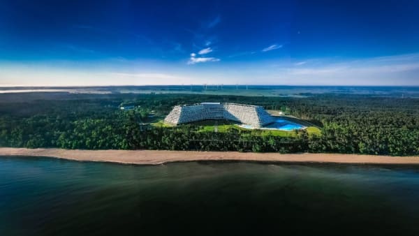 Aerial view of a modern curved resort complex tucked in a forest along a sandy beach with a bright blue pool courtyard nearby.