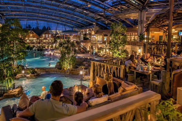 Indoor resort atrium with a central pool, tropical plants, and diners at wooden tables along a balcony grocery.