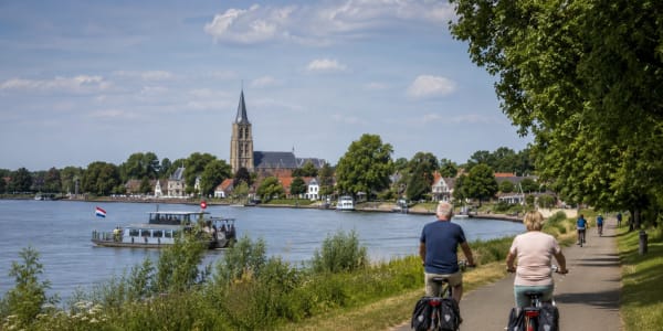 Cyclists ride along a riverside path with a town and tall church spire across the water under a blue sky.