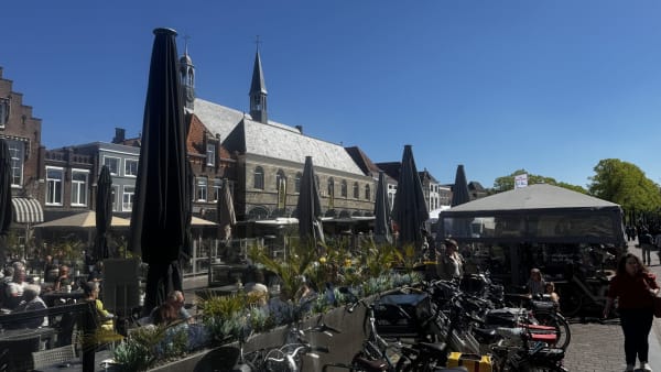 Civil plaza with outdoor cafe tables under large closed black umbrellas, historic brick buildings and church steeples in the background, bright blue sky.