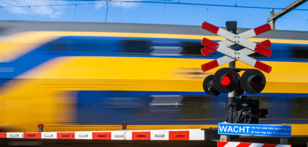 Blurred blue-and-yellow passenger train speeds by at a level crossing; red warning lights are illuminated and a blue 'WACHT' sign is visible.