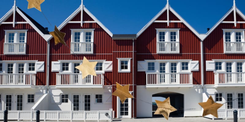 Row of red wooden houses with white trim and balconies under a blue sky, gold star garlands in the foreground.