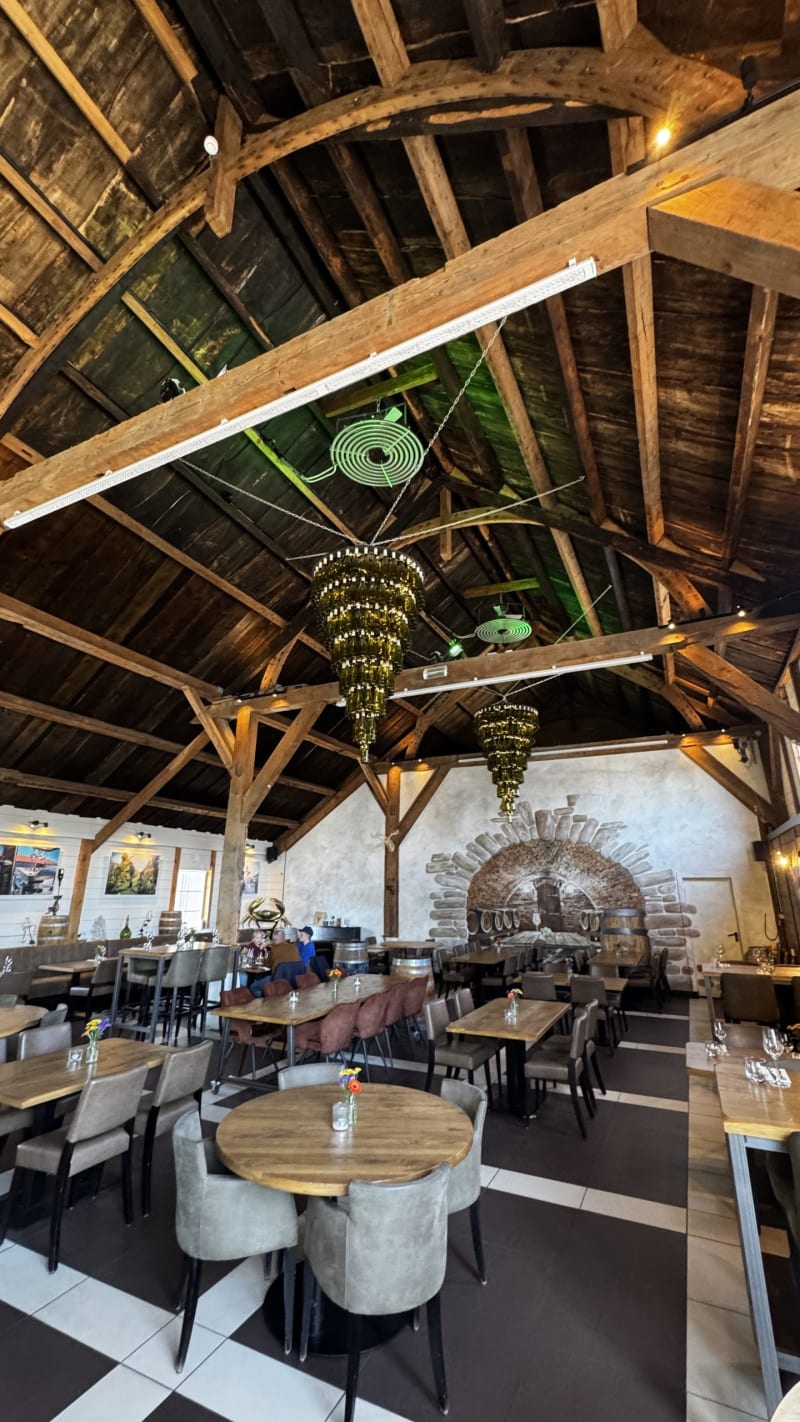 Rustic restaurant interior with exposed wooden beams, vaulted ceiling, and many tables set for dining under warm lighting.