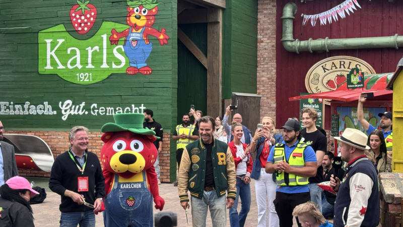 Crowd at a festival in front of a green wall mural advertising Karl's 1921, with a cartoon strawberry character and a bear mascot in blue overalls nearby.