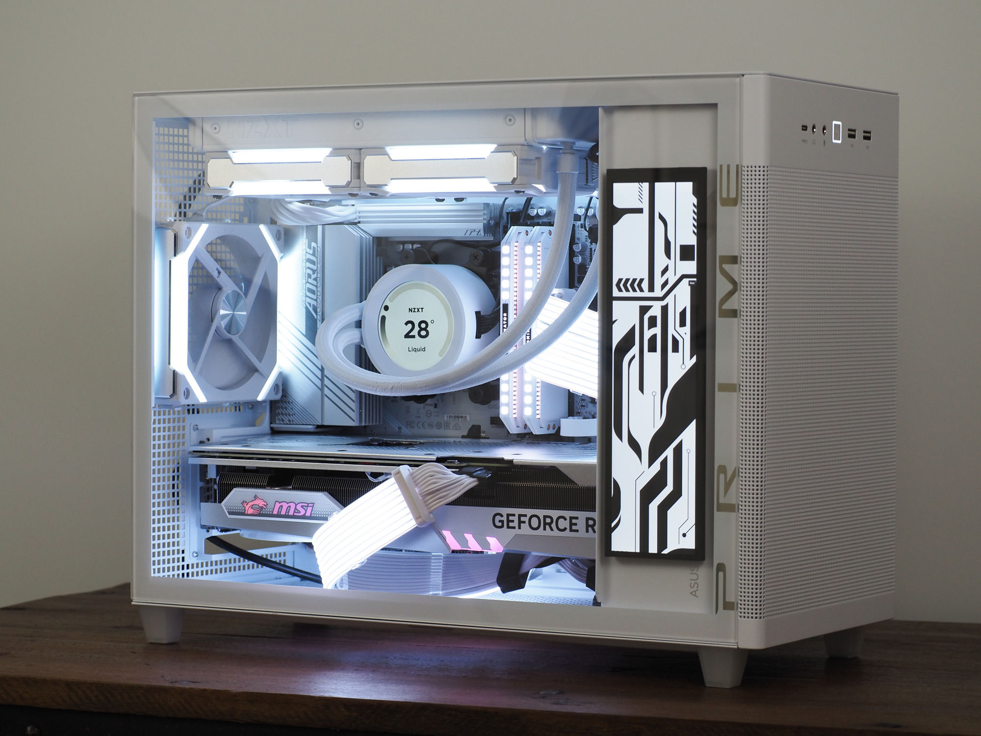 A white Optimist build — NZXT Kraken display reading 28°, a “PRIME” circuit-art side panel and MSI GeForce GPU, on a wooden desk