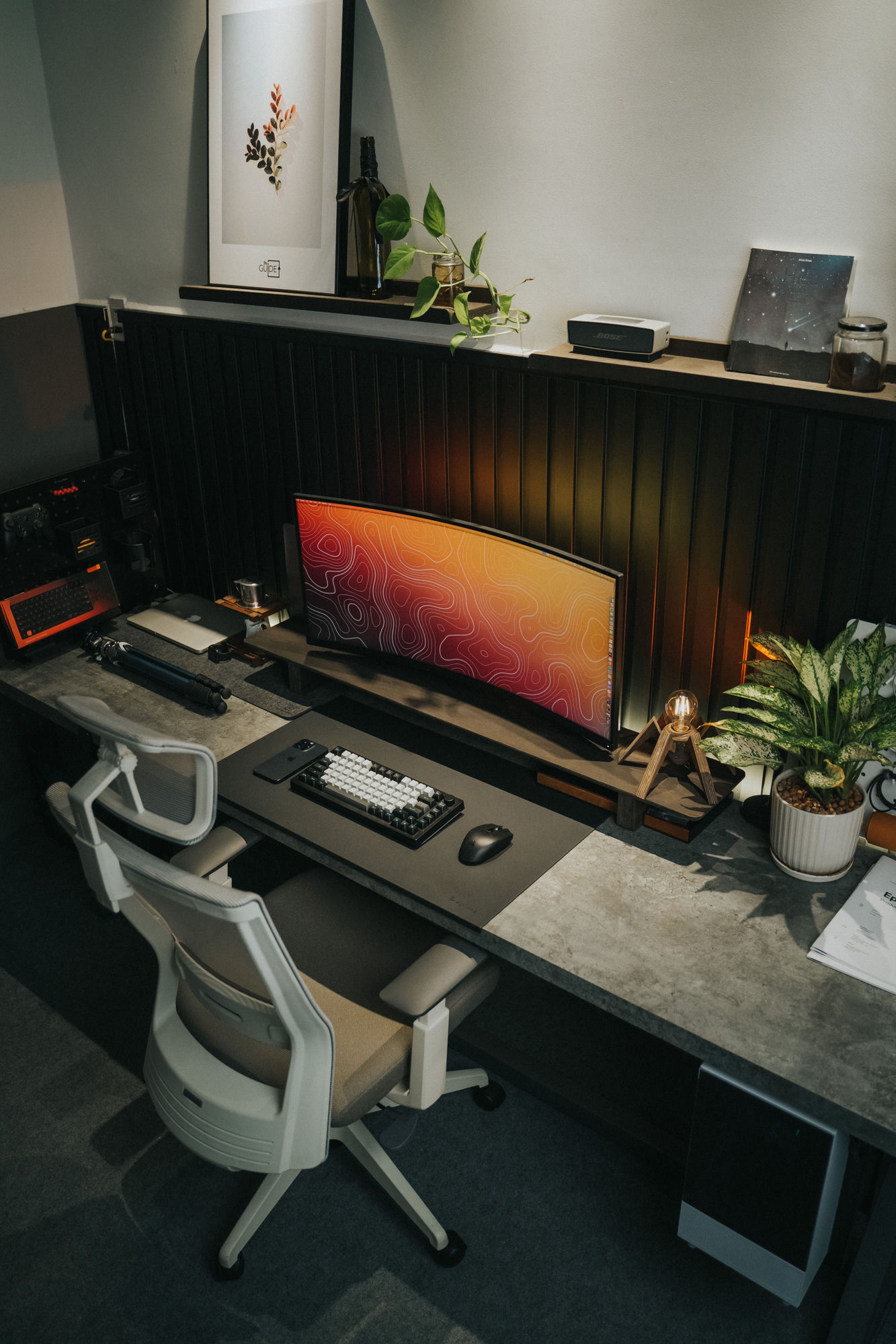 A calm, minimal desk — a curved ultrawide monitor with a topographic wallpaper, a mechanical keyboard, warm bulb lamp and plants against dark wood paneling
