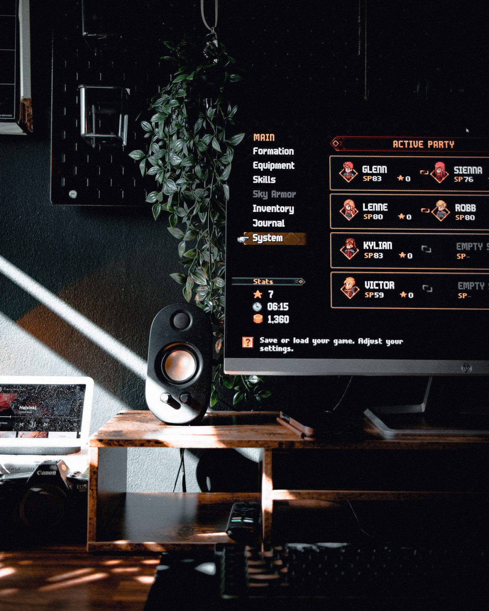 A warm, sunlit desk — a monitor running a pixel-art RPG menu, a speaker, a hanging plant, a laptop and a camera