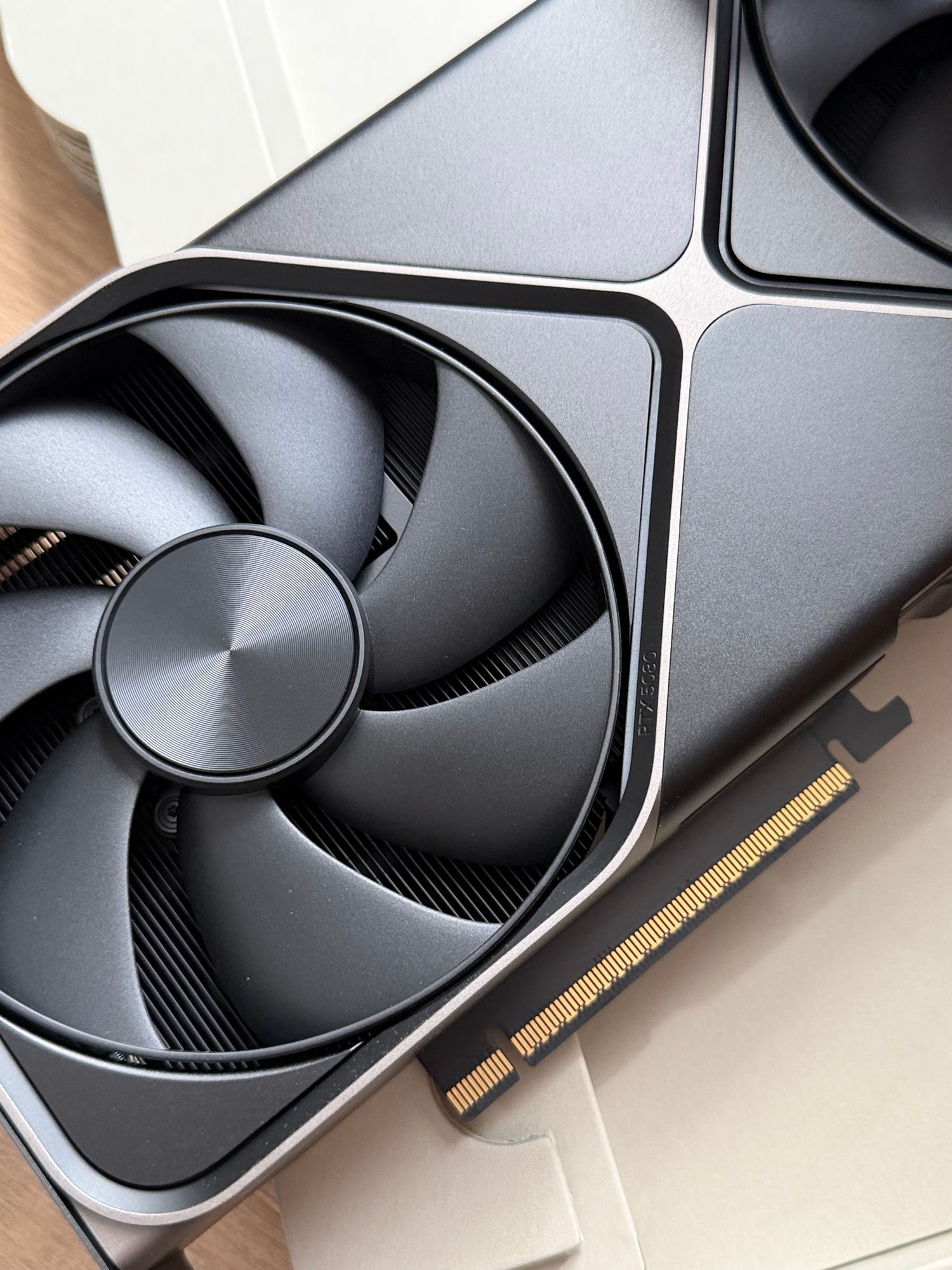 A close-up of a modern RTX 5080 graphics card fan and its gold PCIe edge connector on a light desk