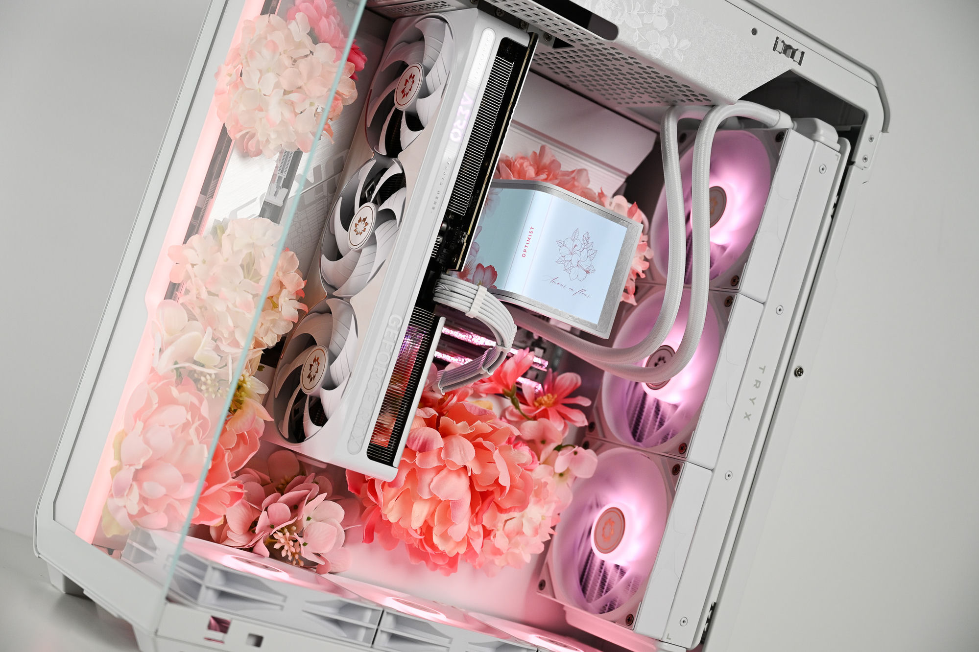 The Fleur — a white TRYX tower shot front-on against seamless white, the whole machine in view: an etched OPTIMIST top panel, an interior packed with pink and coral blooms, a white AERO GeForce RTX card, and the “Amour en fleur” pump screen glowing above pink-lit fans