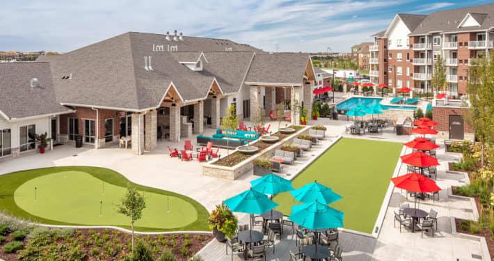 Aerial view of residential community clubhouse with putting green, blue umbrellas, red patio umbrellas, pools, and multi-story apartment buildings.