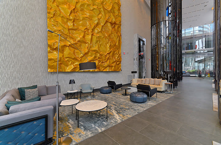 Modern lobby lounge with gray sofa, round tables, blue accents, and large gold abstract wall art beneath floor-to-ceiling windows.
