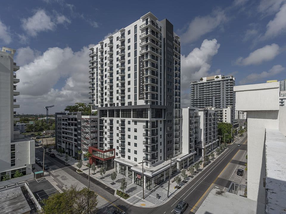 White and dark multi-story residential building with balconies, ground-floor retail, and surrounding urban streetscape under blue sky.