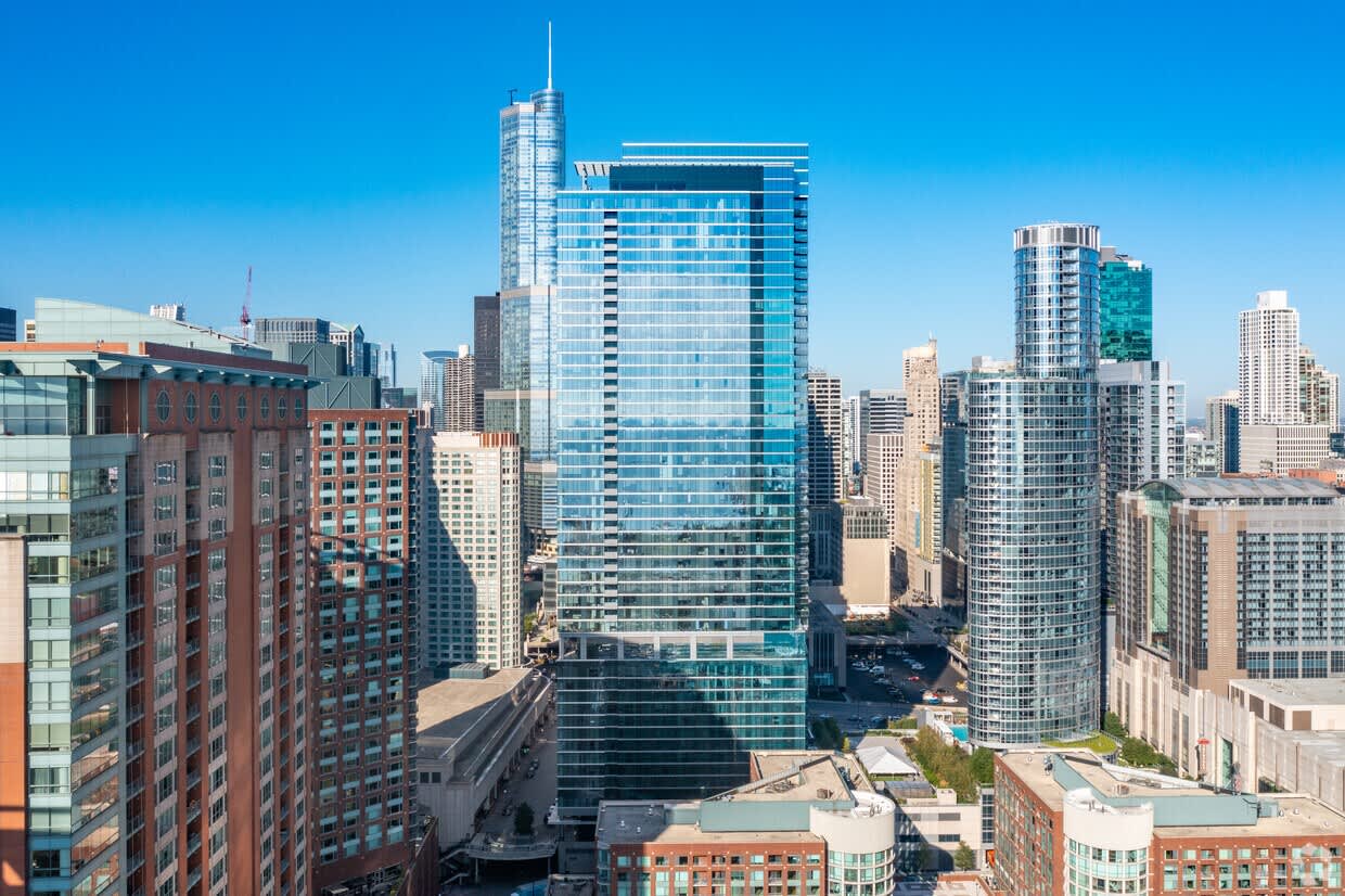 High-rise apartment building with glass facade overlooking Chicago downtown, surrounded by mixed-height buildings under clear blue sky.