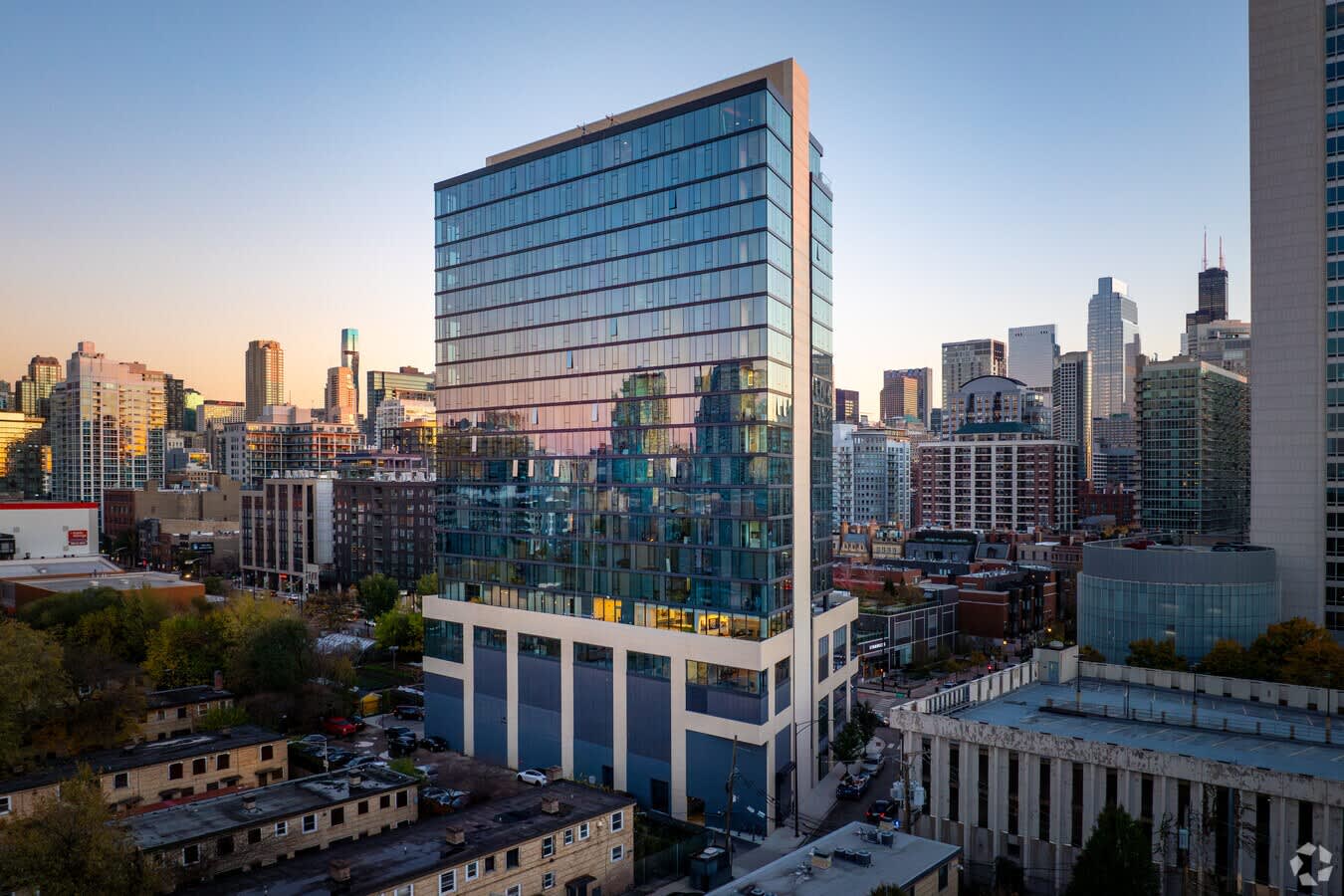 Modern glass and steel residential tower with blue-tinted facade overlooking Chicago skyline at dusk, surrounded by downtown buildings.