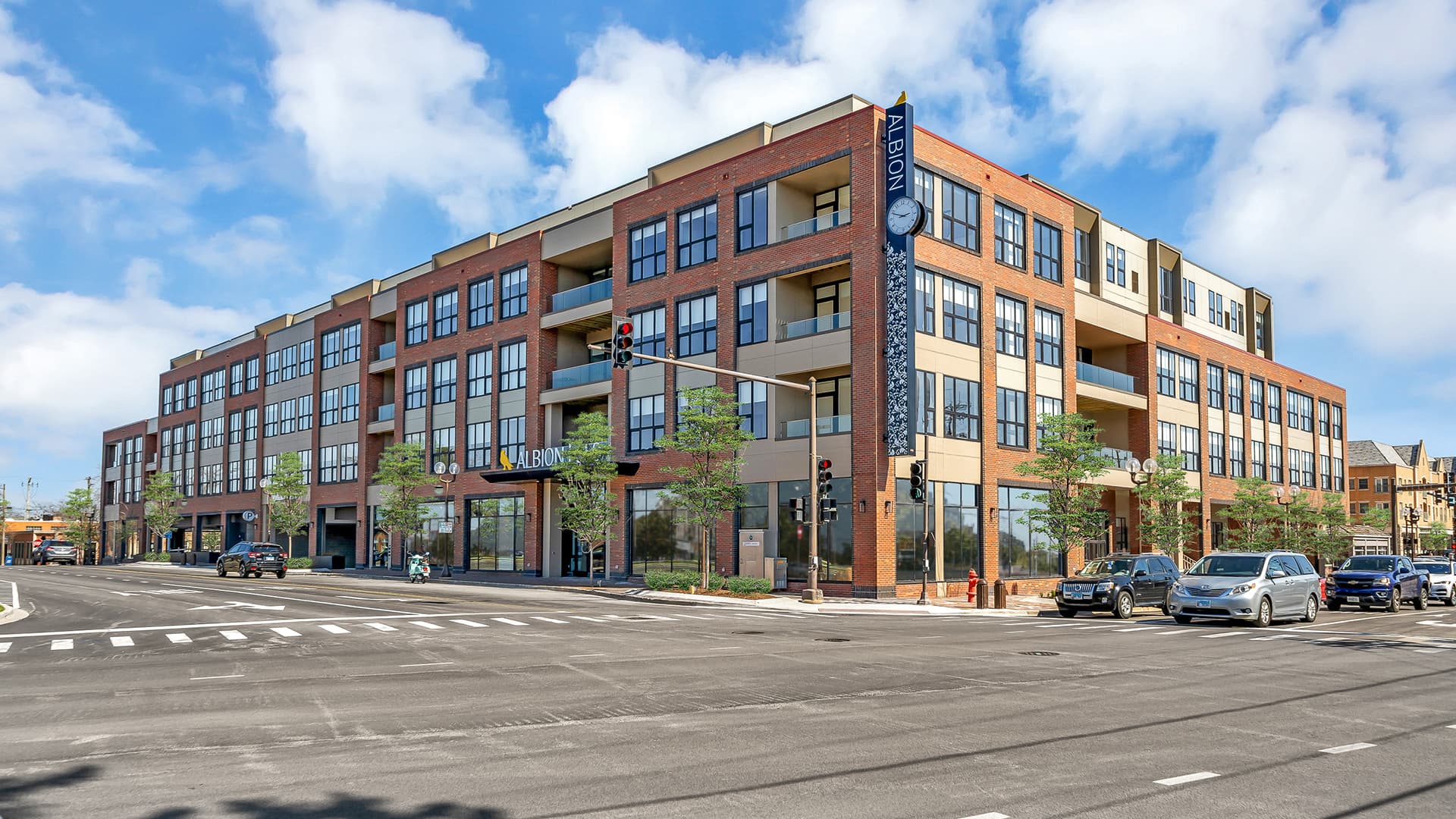 Modern brick and tan residential building with multiple balconies, large windows, and ground-floor retail space on a tree-lined street.