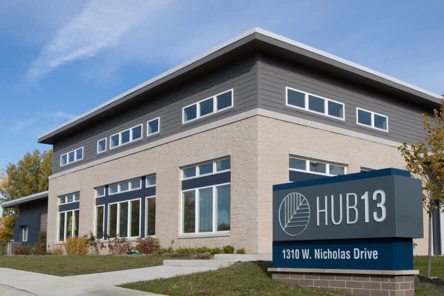 Modern two-story building with cream brick and dark gray upper level, multiple large windows, and Hub13 signage at 1310 W. Nicholas Drive.
