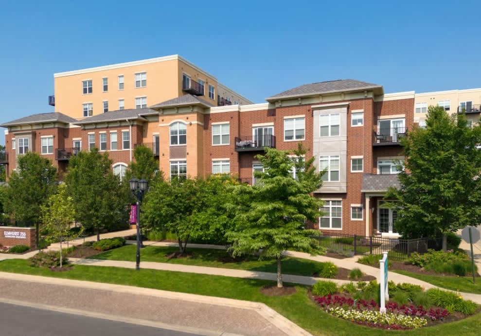 Multi-story brick apartment complex with mature trees, green lawn, and flowering landscaping in front.