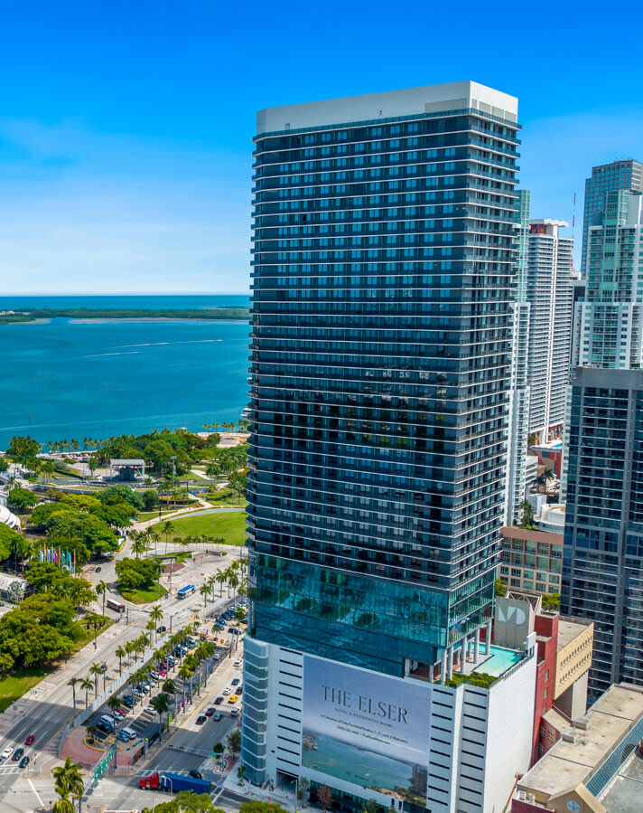 Modern high-rise residential tower with blue-tinted glass facade overlooking turquoise ocean, palm trees, and waterfront park in Miami.
