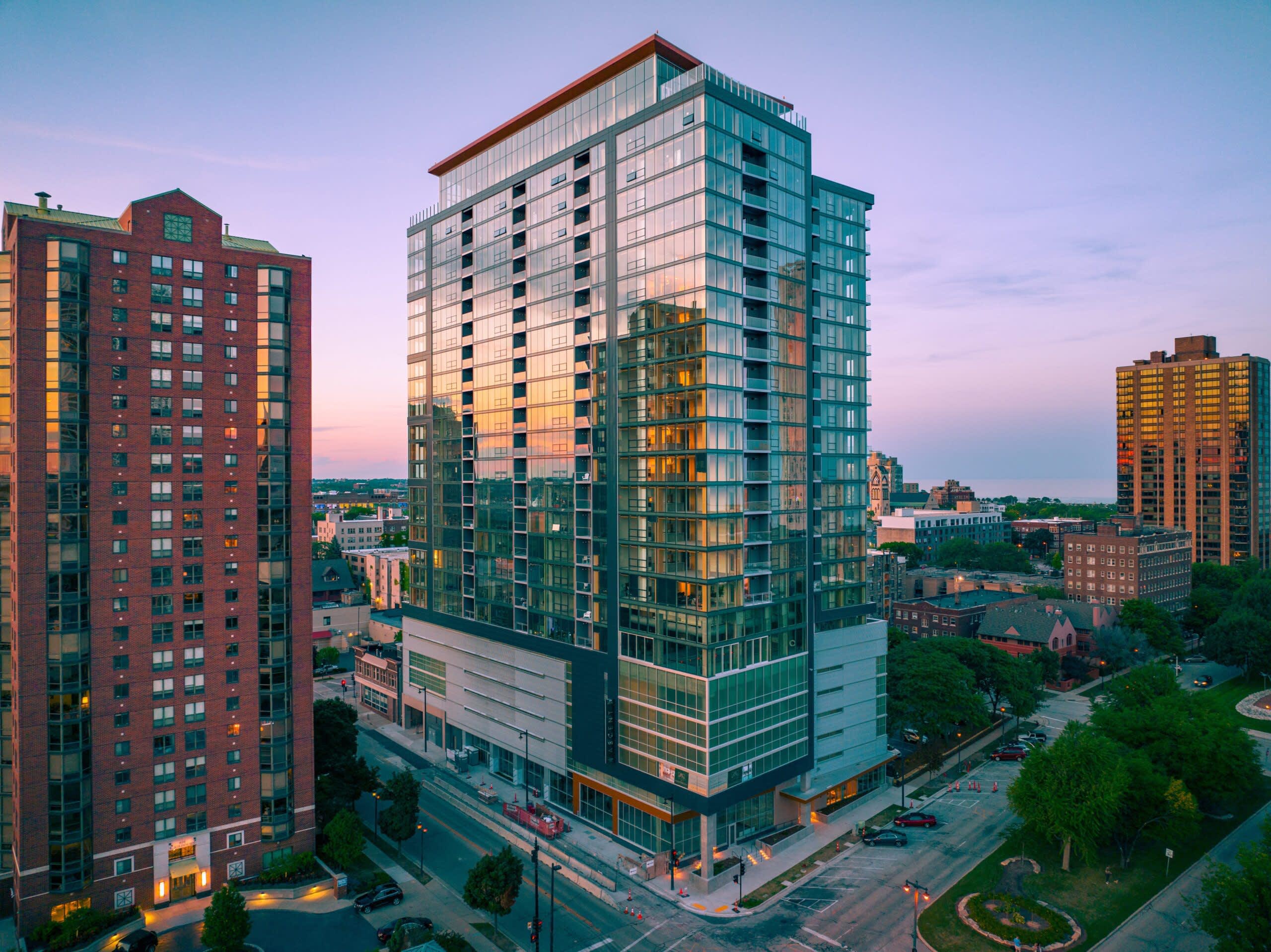 Modern high-rise residential building with green-tinted glass facade and illuminated windows at dusk, surrounded by older brick buildings in an urban setting.